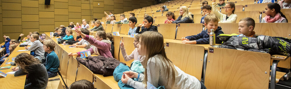 Zahlreiche Kinder hören in einem Stufenhörsaal interessiert einer Vorlesung zu
